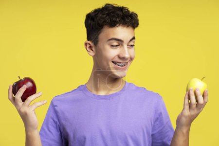 Foto de Guapo y sonriente chico excitado, adolescente con aparatos ortopédicos sosteniendo manzanas recogiendo, mirando, de pie aislado sobre un fondo amarillo. Concepto de comida saludable, fruta - Imagen libre de derechos