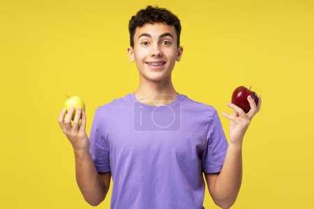 Téléchargez les photos : Garçon excité et souriant portant une tenue élégante tenant une pomme rouge et jaune regardant la caméra. Adolescent attrayant debout isolé sur fond jaune. Concept de vitamines, fruits - en image libre de droit