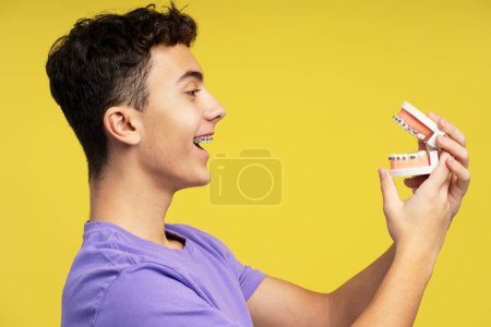 Foto de Sonriente joven atractivo, adolescente con frenos que sostienen la mandíbula ortodóncica, mirándola de pie aislada sobre un fondo amarillo. Concepto de odontología, tratamiento - Imagen libre de derechos