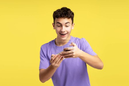 Téléchargez les photos : Un adolescent excité, souriant et séduisant tenant son téléphone portable, utilisant une application mobile, jouant à un jeu, isolé sur fond jaune. Concept de divertissement - en image libre de droit