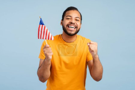 Foto de Feliz hombre afroamericano con frenos dentales, sosteniendo la bandera estadounidense, haciendo un gesto de "sí" con los puños cerrados, y mirando a la cámara, aislado sobre fondo azul. Esperando concepto de resultados electorales - Imagen libre de derechos