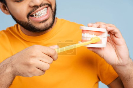 Portrait of a smiling African American man with orthodontic braces, cleaning a plastic jaw model using a toothbrush, isolated on a blue background. Dental care concept