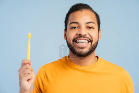 Téléchargez les photos : Portrait d'un homme afro-américain joyeux avec des bretelles, tenant une brosse à dents et regardant la caméra, isolé sur un fond bleu. Concept d'hygiène dentaire - en image libre de droit