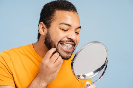 Photo for Portrait of an attractive African American man with orthodontic braces, examining his teeth while looking in a mirror, isolated on a blue background. Dental care concept - Royalty Free Image