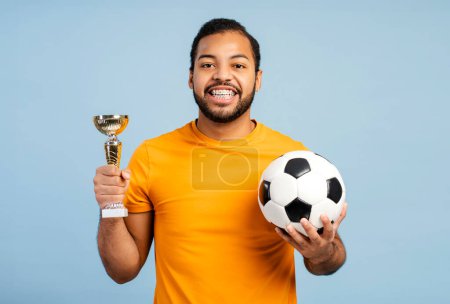 Photo for Portrait of an overjoyed African American athlete with braces, celebrating victory, holding a gold championship cup and soccer ball, looking at the camera, isolated on a blue background - Royalty Free Image