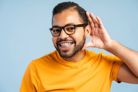 Foto de Retrato foto de un hombre afroamericano sonriente con frenos dentales, usando gafas, haciendo un gesto de escucha como si oyera secretos y mirando a la cámara, aislado sobre un fondo azul - Imagen libre de derechos