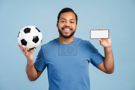 Téléchargez les photos : Joyeux homme afro-américain avec bretelles, tenant un ballon de football et un téléphone portable, montrant l'écran vide avec sa victoire de pari, regardant la caméra isolée sur un fond bleu. Concept de maquette - en image libre de droit