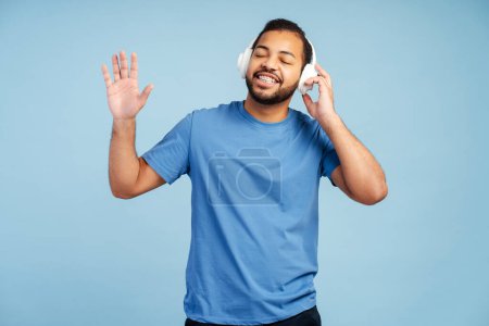 Photo for Cheerful attractive African American man with braces, wearing a headphones, dancing and enjoying music with eyes closed, isolated on a blue background - Royalty Free Image