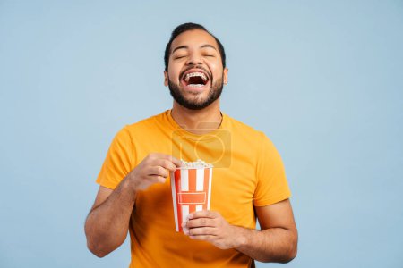 Photo for Overjoyed african american man holding bucket with popcorn, eating snacks, laughing, watching movie isolated on blue background. Cinema, advertisement concept - Royalty Free Image