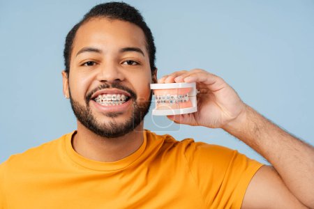 Lächelnder afrikanisch-amerikanischer Mann mit Zahnspange, Zahnform mit Zahnspange in der Hand, lächelnd, isoliert auf blauem Hintergrund. Betonung des kieferorthopädischen Therapiekonzepts 