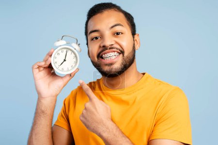 Photo for Happy african-american man with dental braces holding alarm clock and pointing with finger isolated against blue background. Emotional student late to study - Royalty Free Image