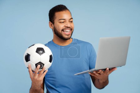 Foto de Retrato de un hombre guapo sonriente sosteniendo la pelota de fútbol, mirando portátil aislado sobre fondo azul. Concepto de hobby, tiempo libre - Imagen libre de derechos
