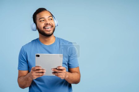 Photo for Smiling handsome man wearing headphones, holding digital tablet, listening to music isolated on blue background. Fun concept, technology - Royalty Free Image
