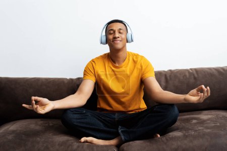 Téléchargez les photos : Homme afro-américain souriant portant un casque sans fil, écoutant de la musique, assis en position lotus, méditant les yeux fermés, assis sur un canapé à la maison. Concept d'équilibre, harmonie, détente - en image libre de droit