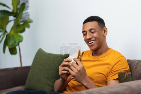 Photo for Portrait of handsome young African American man wearing casual yellow t shirt holding tasty sandwich, eating while sitting on comfortable sofa, copy space. Concept of food, snack, break - Royalty Free Image