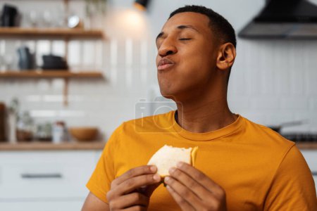 Téléchargez les photos : Portrait d'un bel homme afro-américain heureux tenant un sandwich savoureux, mangeant, appréciant les yeux fermés assis dans la cuisine moderne à la maison. Concept de nourriture, pause - en image libre de droit
