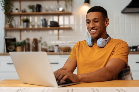 Foto de Sonriendo atractivo hombre afroamericano, programador, estudiante usando el ordenador portátil, usando ropa elegante y auriculares modernos, trabajando desde casa, trabajo remoto en línea. Concepto de educación en línea, tecnología - Imagen libre de derechos
