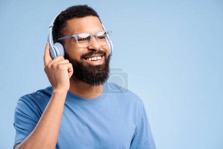 Foto de Atractivo hombre afroamericano feliz con gafas con estilo escuchando música en auriculares mirando hacia otro lado espacio de copia aislado sobre fondo azul. Concepto de publicidad, hobby - Imagen libre de derechos