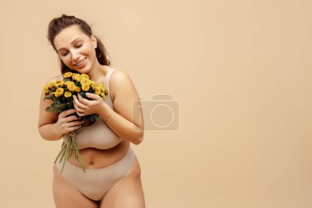 Happy attractive smiling woman wearing stylish lingerie holding bunch of yellow chrysanthemums posing isolated on beige background, copy space. Advertisement concept, International Women's Day