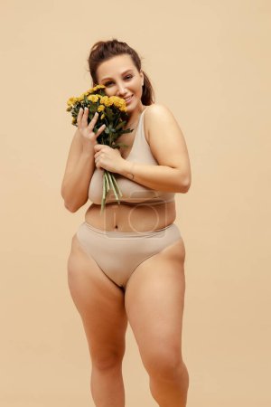 Portrait of cheerful smiling plus size woman wearing stylish sexy lingerie, holding chrysanthemum bouquet, looking at camera standing isolated on beige background. Concept of March 8