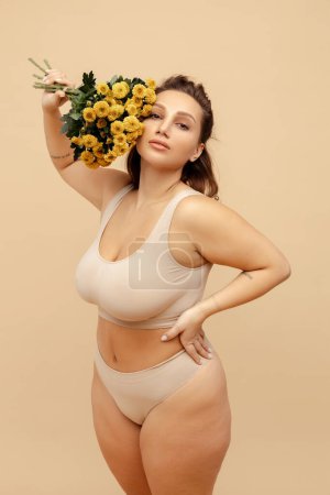 Portrait of attractive body positive woman wearing stylish sexy lingerie, holding yellow chrysanthemum bouquet, standing isolated on beige background. International women's day concept