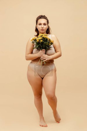 Portrait of body positive woman wearing stylish sexy lingerie, holding bouquet of chrysanthemums, looking at camera standing isolated on beige background. International women's day concept, 8 March
