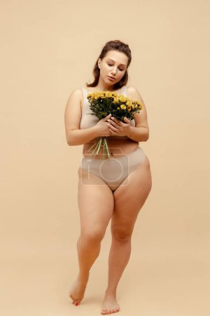 Portrait of beautiful smiling body positive woman wearing stylish sexy lingerie, holding chrysanthemum bouquet, standing isolated on beige background. International women's day concept