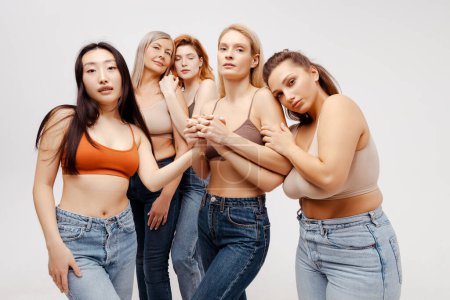 Photo for Modern diverse, multiracial group of women wearing sexy bras and jeans hugging, looking at camera. Fashion beautiful models posing in studio, standing isolated on white background. Support concept - Royalty Free Image