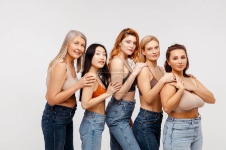 Photo for Modern diverse, multiracial group of women wearing sexy bras and jeans hugging, looking at camera. Fashion beautiful models posing in studio, standing isolated on white background. Support concept - Royalty Free Image
