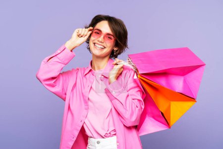 Photo for A beautiful, smiling woman in sunglasses and pink garments, holding several paper shopping bags, standing isolated on a lilac backdrop, looking directly at the camera. Shopping concept - Royalty Free Image