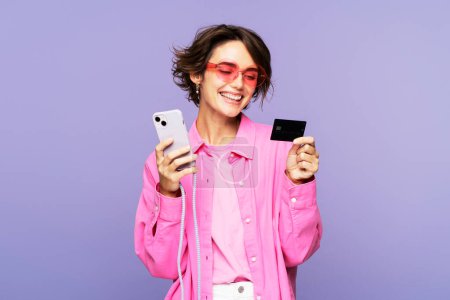Photo for Portrait of a charming woman, wearing sunglasses, holding a smartphone and credit card for shopping, standing isolated on a lilac background. Online shopping application concept - Royalty Free Image