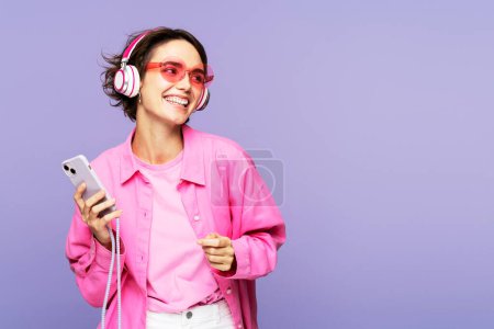 Téléchargez les photos : Photo de portrait d'une femme séduisante, portant des lunettes de soleil et écoutant de la musique dans un casque, tenant son téléphone portable, posant isolée sur un fond lilas et regardant ailleurs. Affiche concept de publicité - en image libre de droit