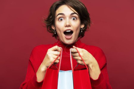 Photo for Portrait photo of an excited, happy woman with wide open mouth holding paper shopping bags with gifts, posing isolated on a red background, and looking at the camera. Sales concept - Royalty Free Image