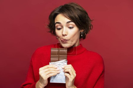 Téléchargez les photos : Portrait d'une femme charmante et drôle tenant une barre de chocolat, le désirant, et léchant ses lèvres, debout seul isolé sur une toile de fond rouge tout en regardant le dessert - en image libre de droit