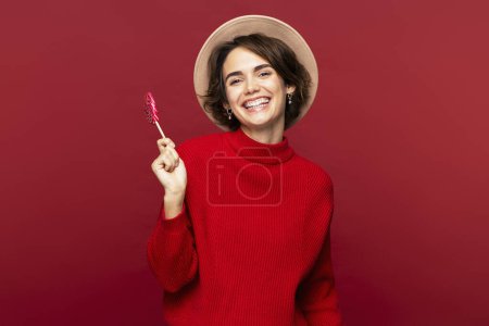 Téléchargez les photos : Photo de portrait d'une femme joyeuse et séduisante portant un chapeau élégant et tenant une sucette douce, posant isolée sur un fond rouge et regardant la caméra - en image libre de droit