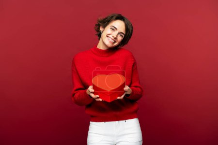 Foto de Fotografía de una mujer feliz y encantadora, sonriendo mientras sostiene una caja de regalo en forma de corazón, aislada sobre un fondo rojo, mirando a la cámara - Imagen libre de derechos