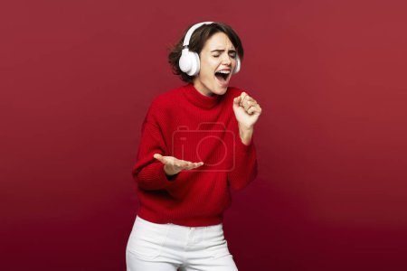 Photo for A portrait of a cheerful, attractive woman with headphones on, listening music, gesticulating and pretending singing into a microphone, isolated on a red background - Royalty Free Image
