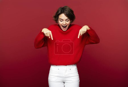 Téléchargez les photos : Photo de portrait d'une femme excitée et séduisante pointant vers le bas avec ses index, démontrant un jean blanc élégant, isolé sur un fond rouge. Concept de mode - en image libre de droit
