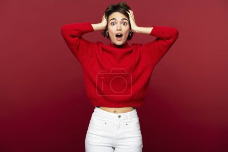 Photo for Portrait of an amazed beautiful woman, emotional female, holding her head with her hands, posing isolated on a red background and looking into the camera - Royalty Free Image