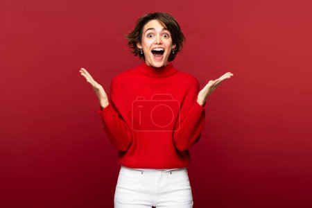 Photo for Attractive young happy woman in a portrait, looking surprised and gesticulating with wide open mouth dramatically, isolated on a red backdrop, looking at the camera - Royalty Free Image