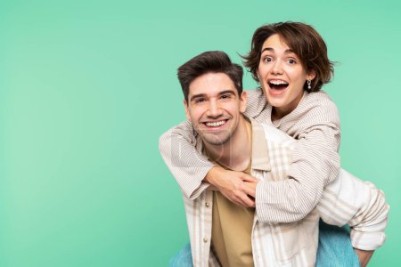Téléchargez les photos : Portrait d'amis joyeux et riants s'amusant, homme portant une femme sur le dos, souriant et posant isolé sur un fond turquoise, regardant la caméra - en image libre de droit