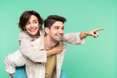 Photo for Attractive, smiling friends in a portrait, where the man holding the woman on his back. She pointing out with her forefinger, both posing on a turquoise background, looking away - Royalty Free Image