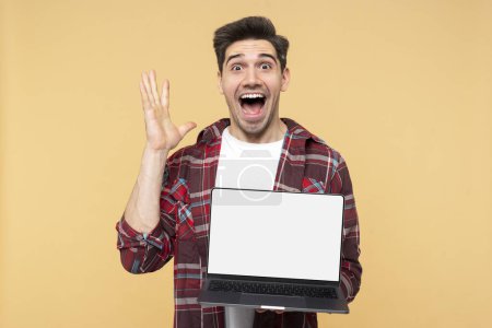 Photo for Portrait photo of a shocked, handsome man gesticulating and shouting, demonstrating a laptop with a blank screen for a promo template, isolated on a yellow background and looking at the camera - Royalty Free Image
