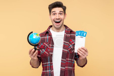 Photo for Portrait photo of an excited man, holding a globe and plane tickets, eyes wide open in surprise, thrilled adventure winner, isolated on a yellow background, looking at the camera - Royalty Free Image