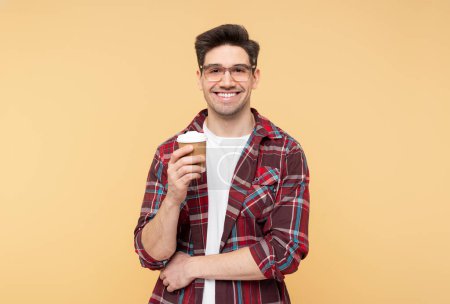 Foto de Foto de retrato de un hombre sonriente y atractivo con gafas, sosteniendo una taza de papel de bebida caliente, posando sobre un fondo amarillo aislado y mirando a la cámara - Imagen libre de derechos