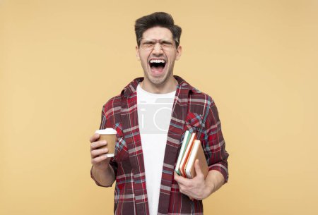 Photo for Portrait of an emotional man in eyeglasses shouting, with eyes closed, holding a paper coffee cup and books, showing morning stress, isolated on a yellow background - Royalty Free Image