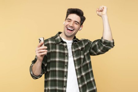 Photo for Smiling man captured in a portrait, holding his mobile phone, dancing, and singing into earbuds, enjoying music, isolated on a yellow background, eyes closed - Royalty Free Image