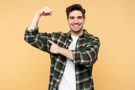 Photo for A portrait shot of a smiling man, showing a muscle gesture, pointing at his biceps with his forefinger, standing before an isolated yellow background and looking at the camera - Royalty Free Image