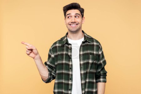 Photo for Portrait photo of a handsome, emotional joyful man with wide smile pointing somewhere with his forefinger, posing against an isolated yellow background and looking away - Royalty Free Image