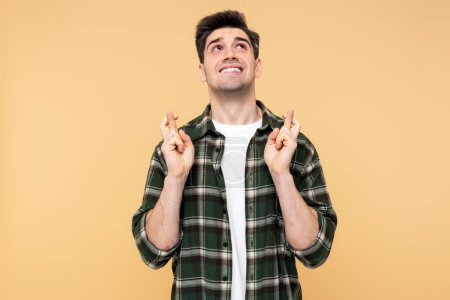 Photo for Portrait of an attractive, hopeful man crossing his fingers in a gesture of hope, biting his lip and praying, posed against an isolated yellow background, looking up - Royalty Free Image